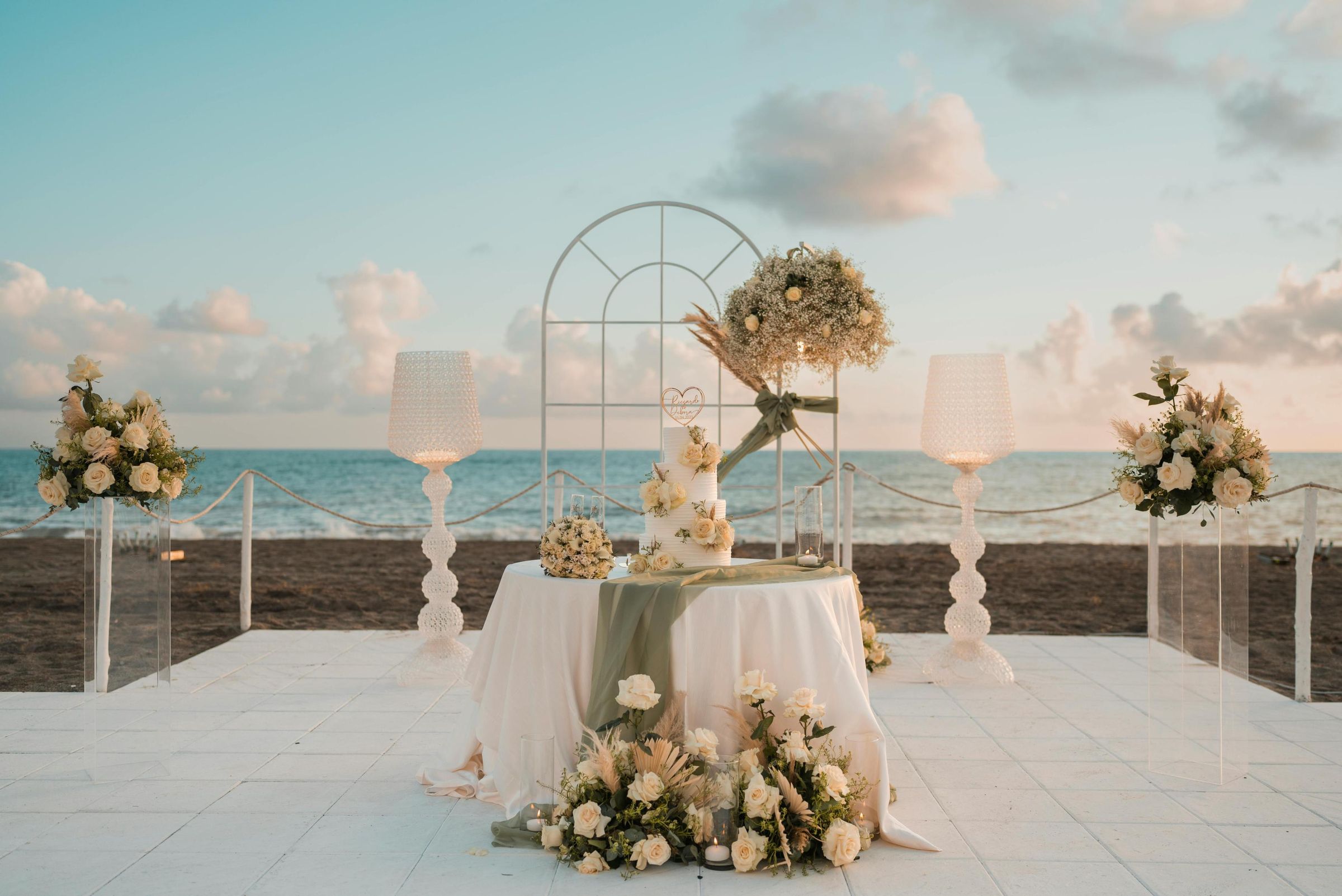 Tiered wedding cake on a draped table framed by a floral arch at a beachfront ceremony at dusk
